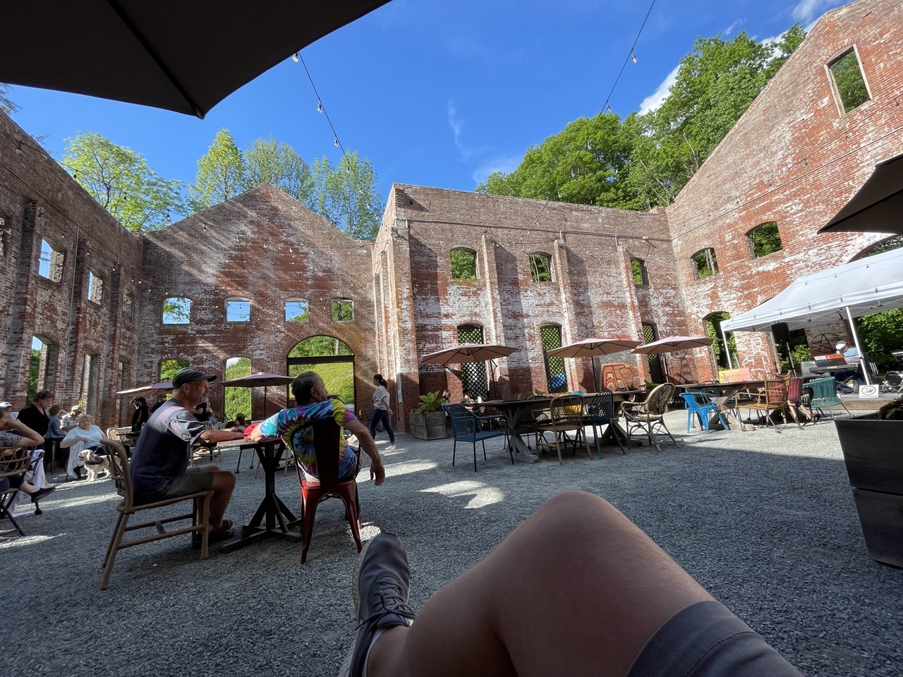 My point of view while sitting inside a hollowed out building turned into a space to eat, drink and listen to music. Blue skies, brick walls, people sitting at tables under umbrellas.