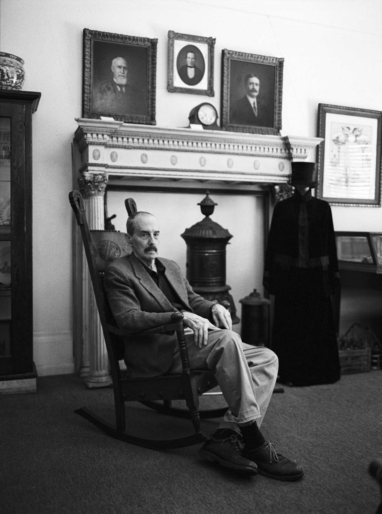S. Robert Powell, Ph.D. at the Carbondale Historical Society sitting in a large rocking chair in front of the faux fireplace with other photos on the wall.