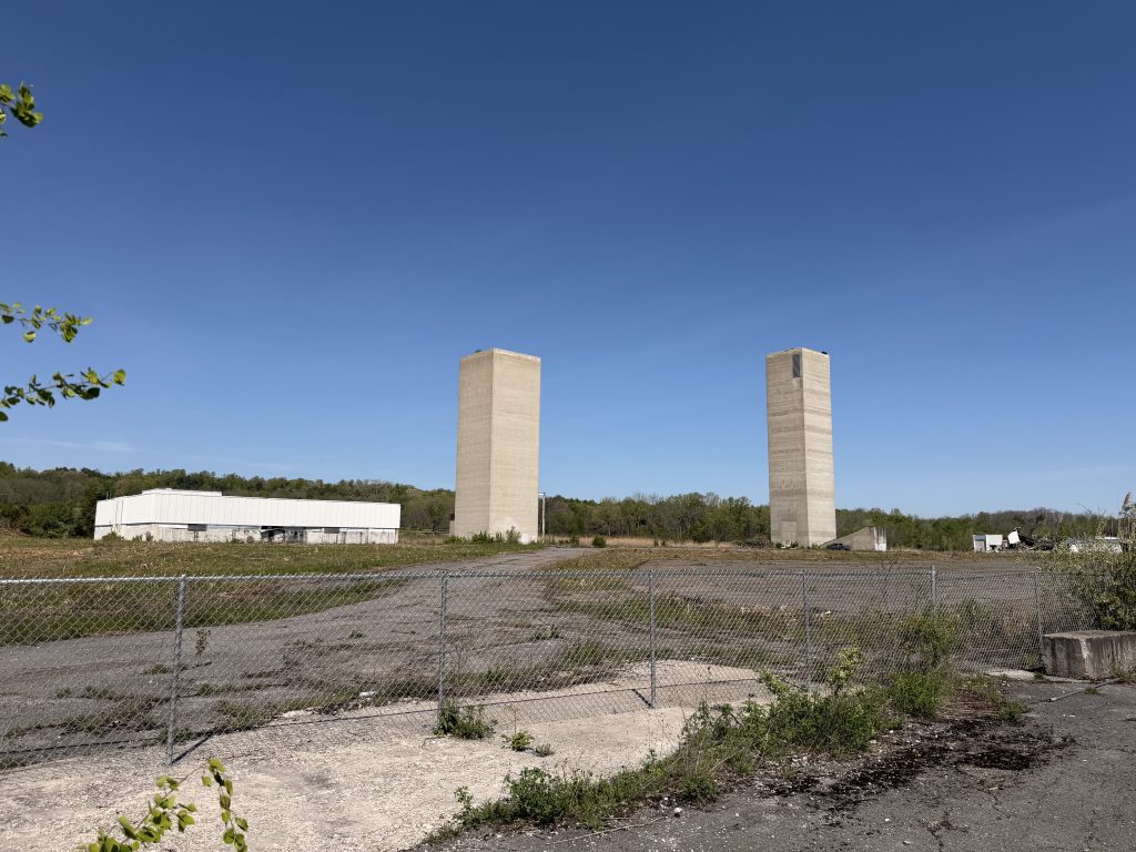 An office building and two stack houses sit in an abandoned parking lot beyond a chainlink fence.