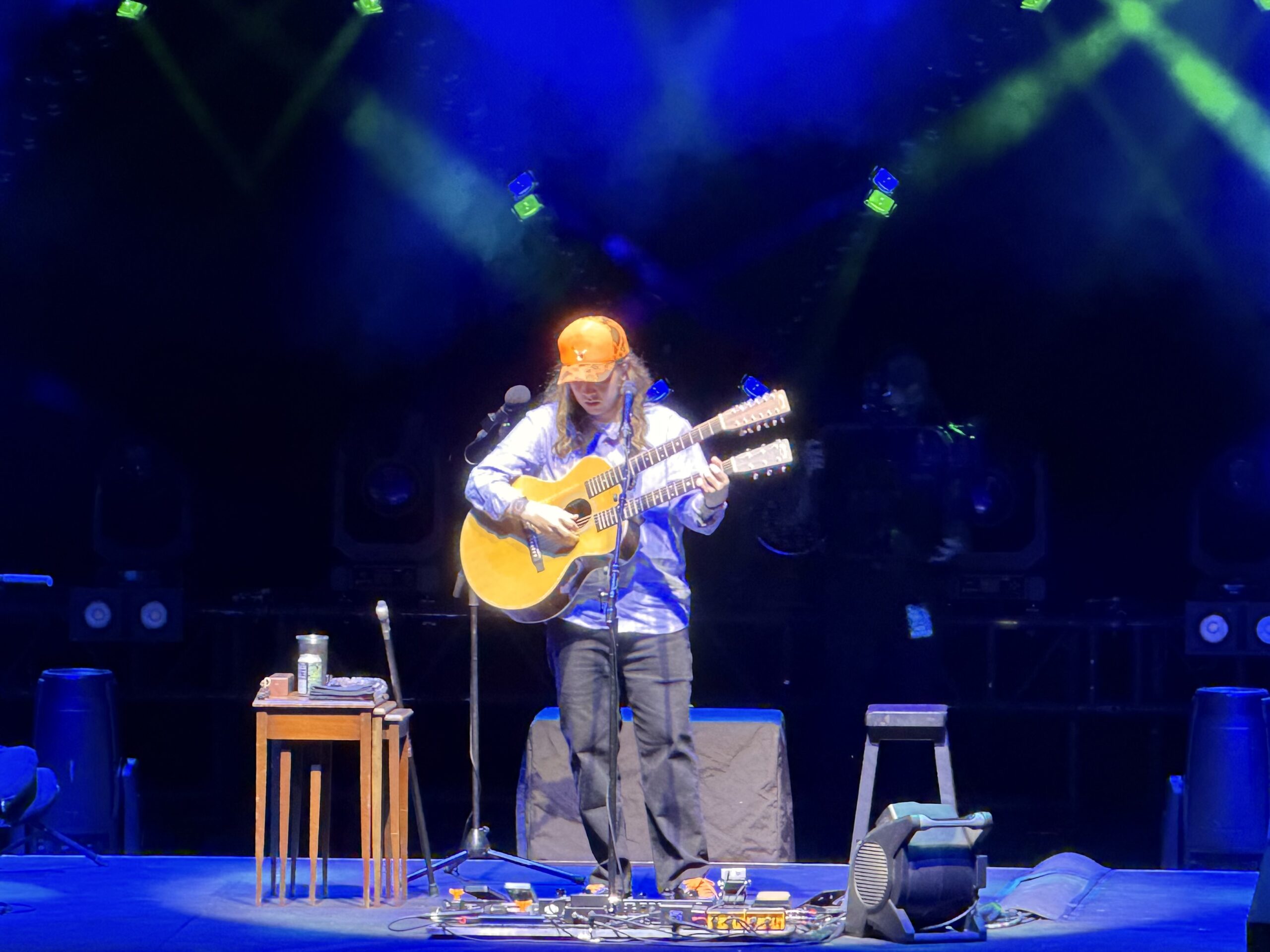 Billy Strings, in an orange cap, plays a double-neck acoustic guitar alone on a stage lit in blue and green.