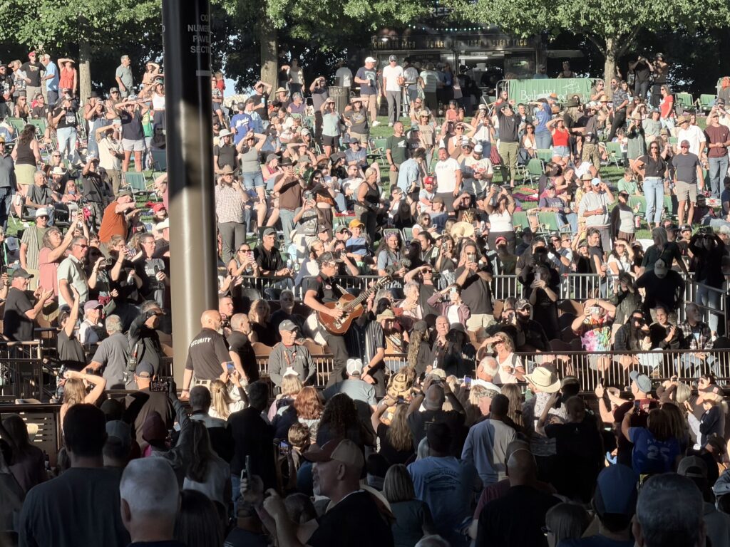 Stephen Wilson Jr. plays an acoustic guitar in the middle of a packed lawn crowd, with fans around him filming on their phones.