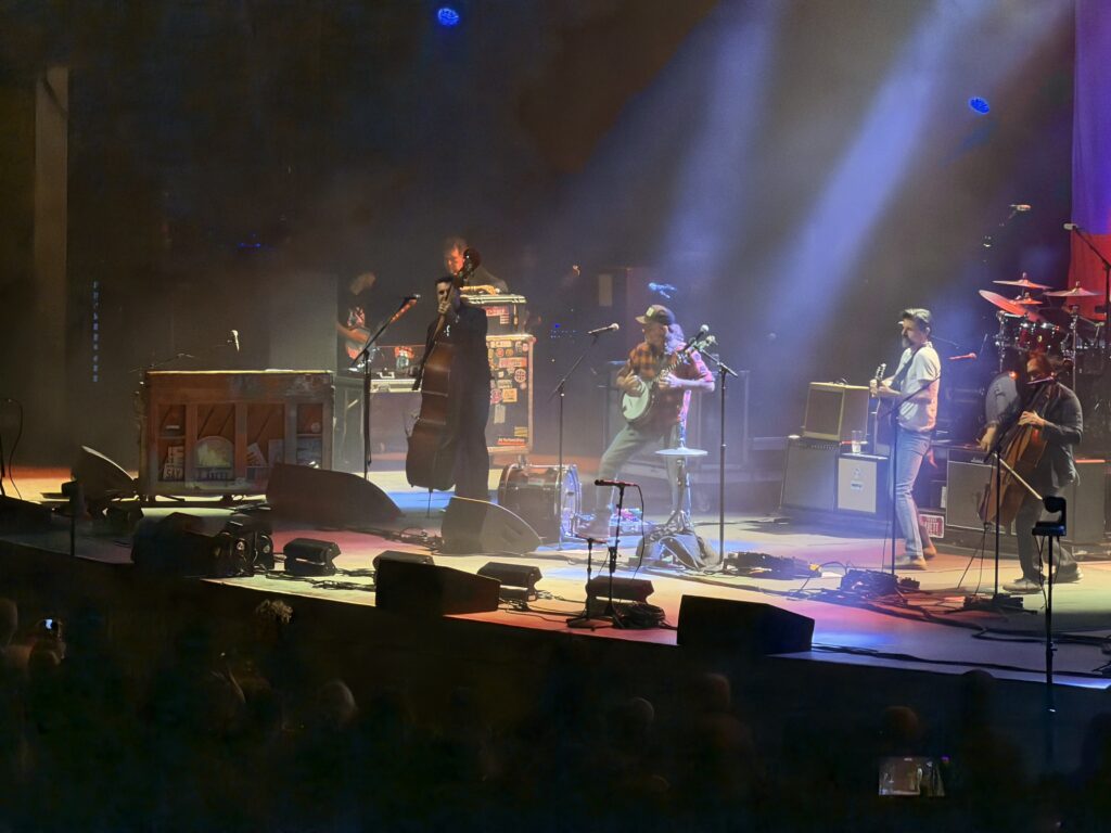 The Avett Brothers on a hazy, spotlit stage with banjo, upright bass, guitar, cello and drums.
